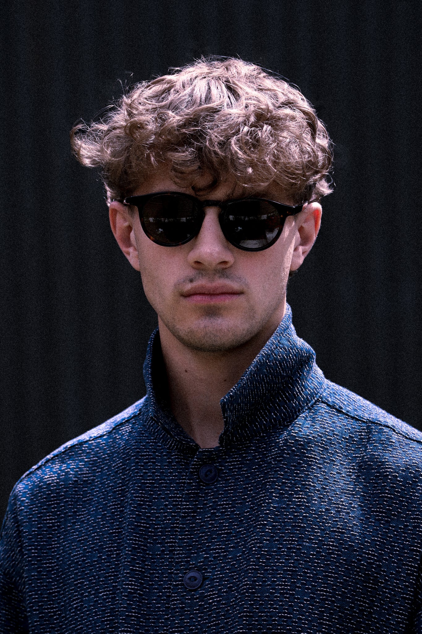 Man wearing sunglasses and a dark blue jacket against a black background